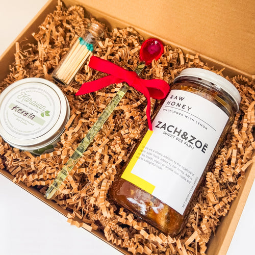 Gift box with jar of honey, decorative glass spoon, small candle, and jar of matches on a light background 